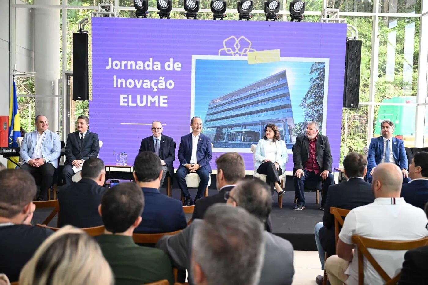 Plateia acompanha mesa de convidados na Jornada de Inovação Elume, com telão roxo ao fundo