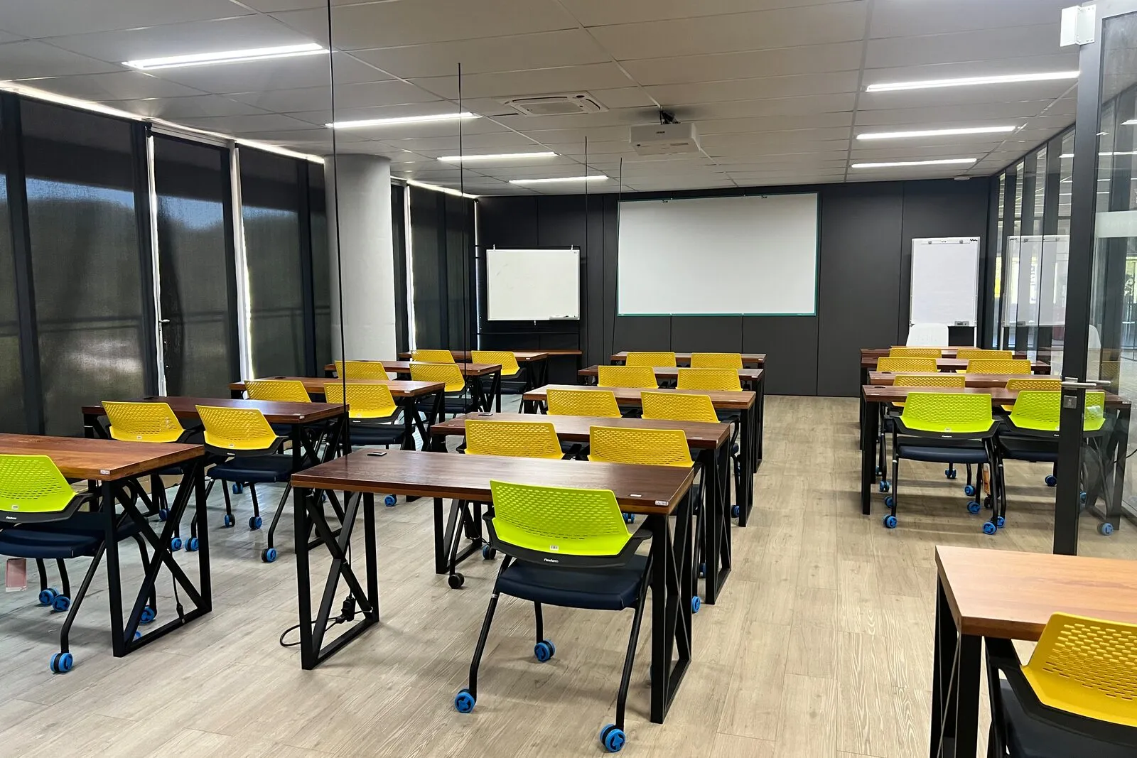 Sala de capacitação do Elume com mesas em fileiras, cadeiras verdes, lousa branca e tela de projeção